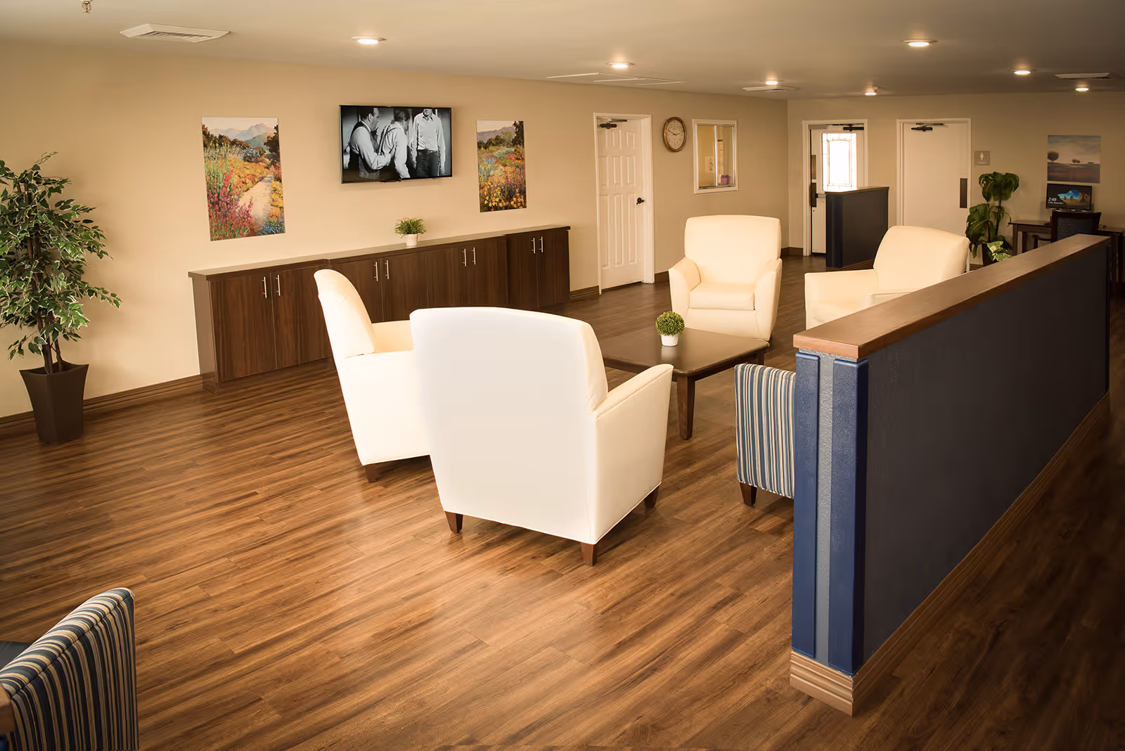 A well-lit lounge area with four cream-colored armchairs arranged around a wooden coffee table with a small plant on it. The room has wood flooring, a dark wooden cabinet against the wall, two landscape paintings, a wall-mounted TV showing a black and white image, a clock, and some plants. There is a blue partition wall on the right side and a door in the background.