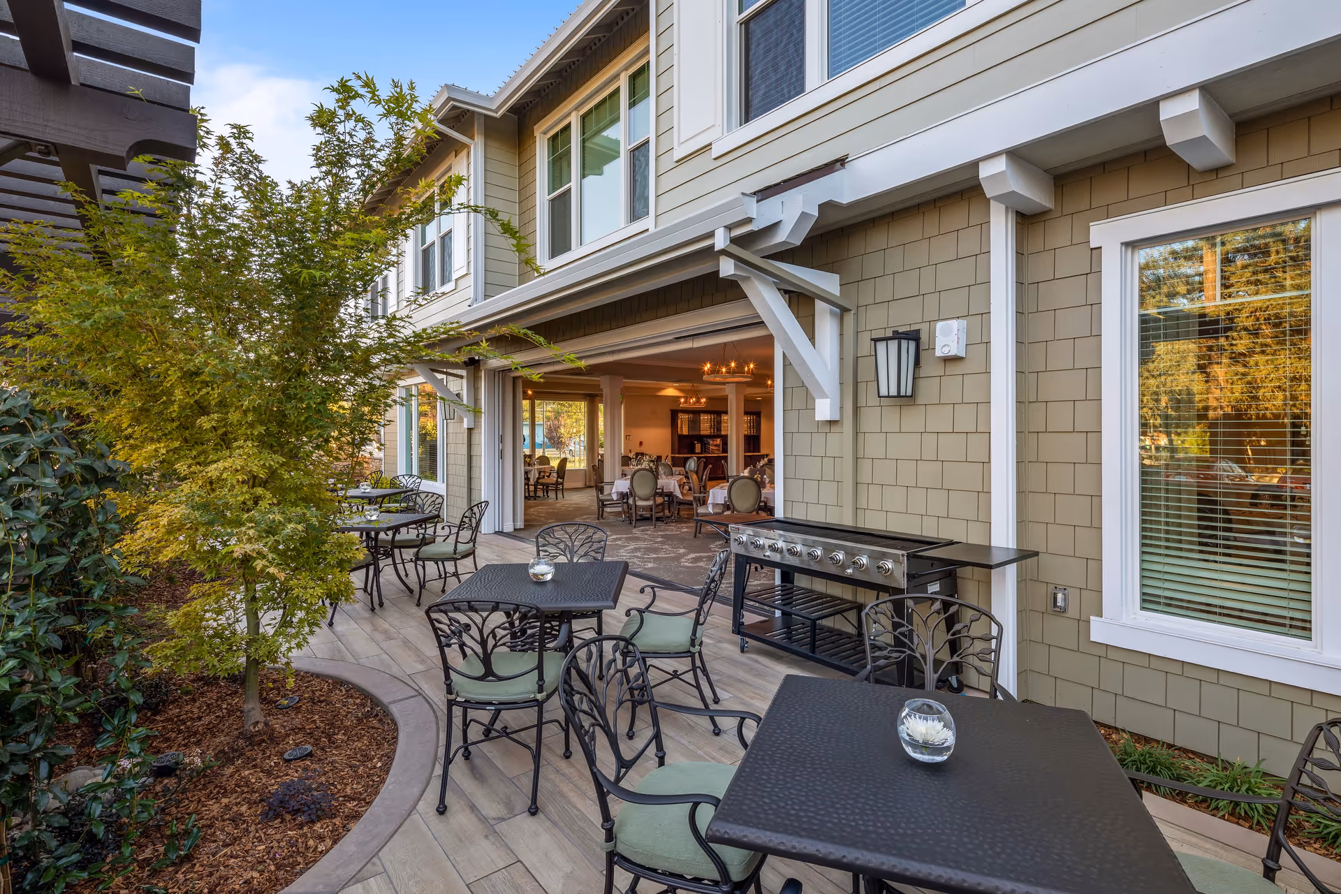 Outdoor patio area at Oakmont of Novato with several black metal tables and chairs with green cushions. A small tree and plants are along the left side, and a large grill is positioned against the building wall. The patio opens into an indoor dining area with tables and chairs visible inside.