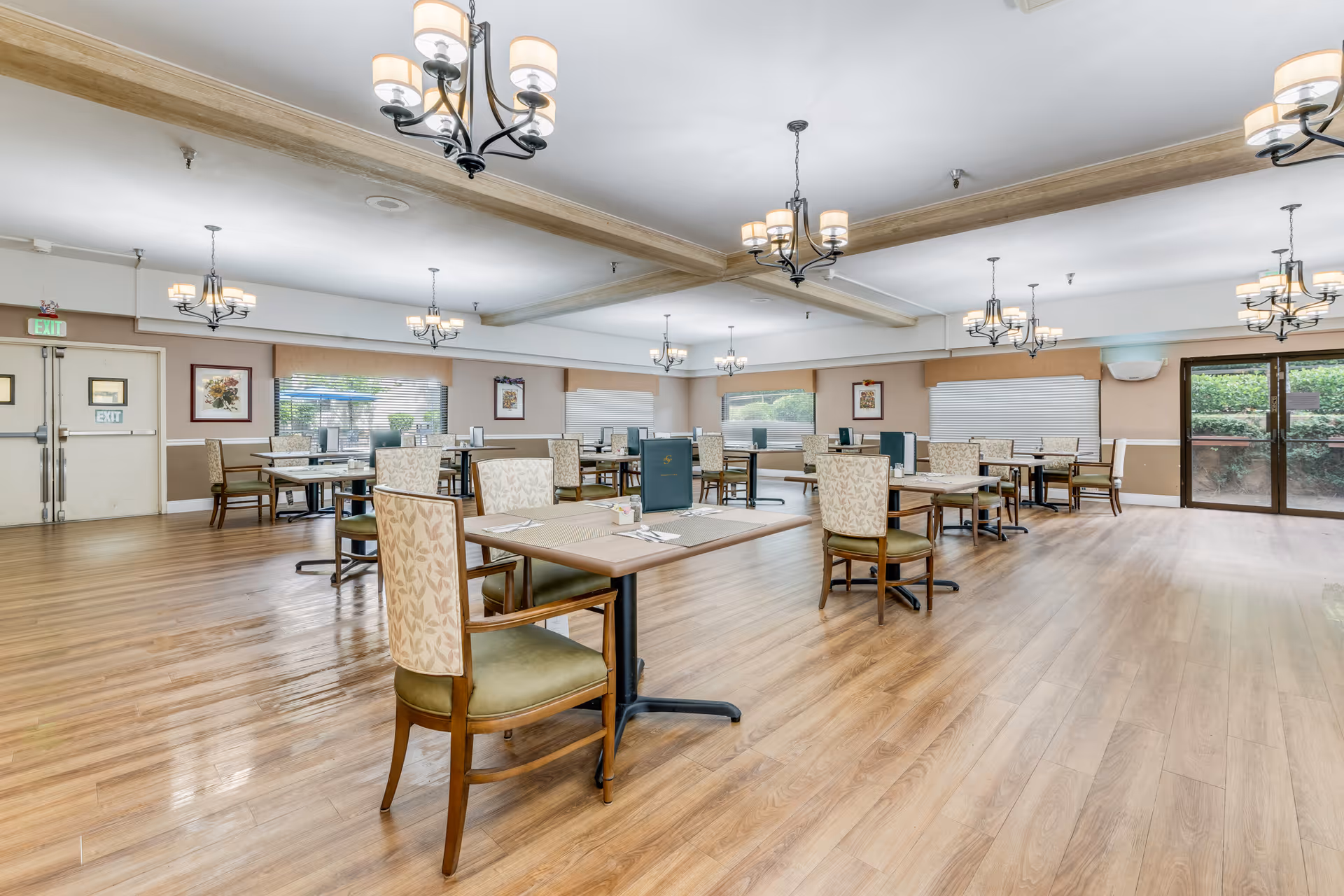 Spacious senior dining room with multiple tables and upholstered chairs, chandeliers, large windows, and wood flooring.