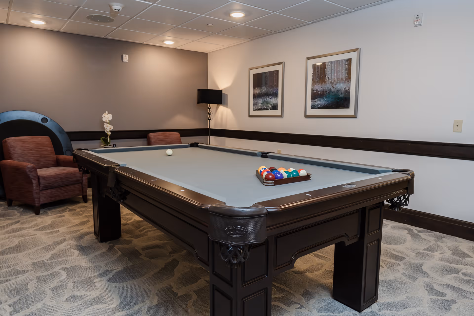 A recreational room with a pool table set up for a game, featuring a rack of colorful billiard balls and a cue ball. The room has two upholstered chairs, a floor lamp, two framed abstract paintings on the wall, and a patterned carpet.
