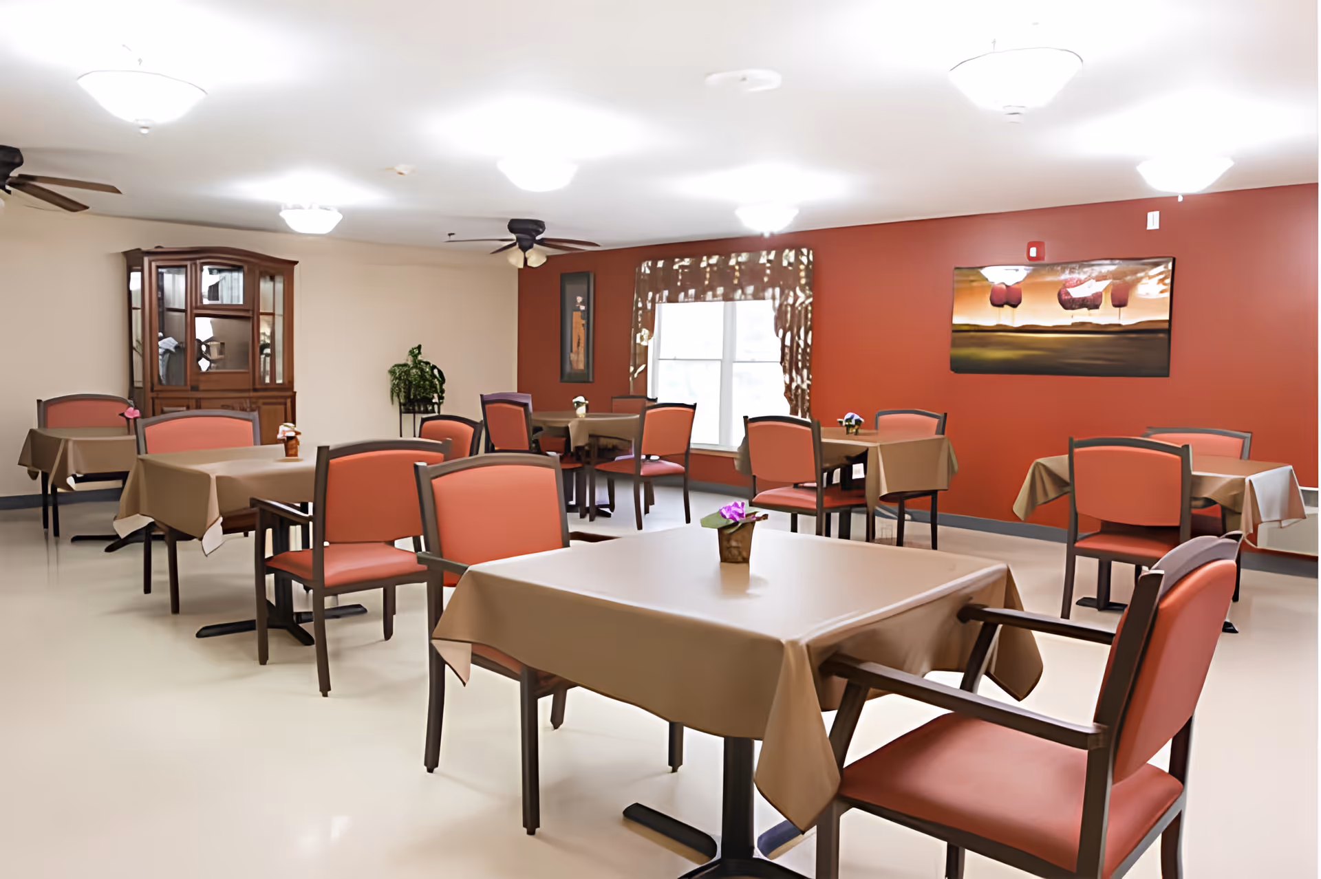 A dining room with multiple tables covered with beige tablecloths and surrounded by chairs with coral-colored cushions. The room has a red accent wall with a window and artwork, ceiling lights, ceiling fans, and a wooden cabinet in the corner.