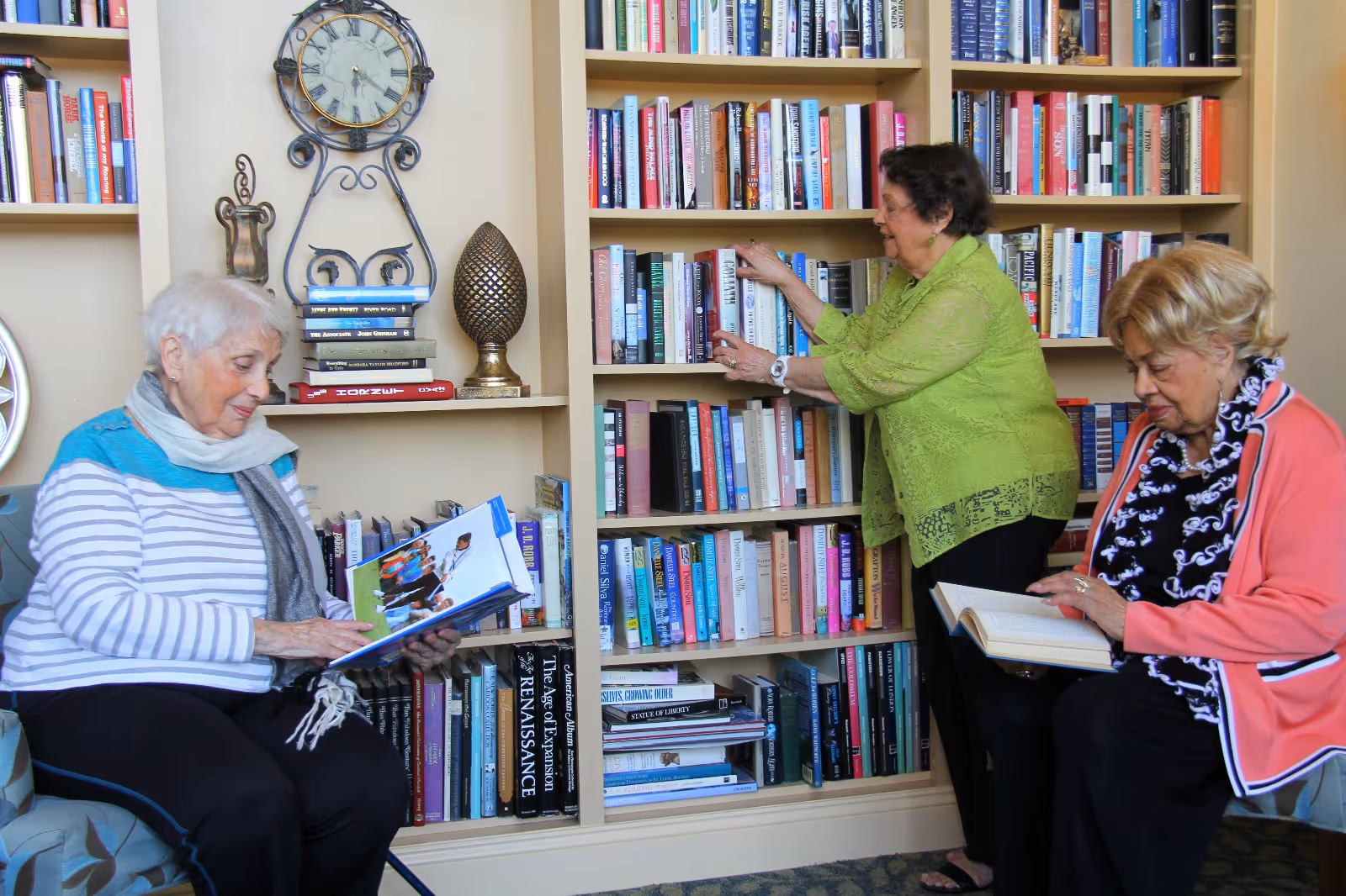 Three elderly women in a library or reading room. One woman in a striped shirt and scarf is sitting and reading a book, another woman in a green blouse is standing and selecting a book from a bookshelf filled with books, and the third woman in a pink jacket is sitting and reading a book. The room has built-in bookshelves with many books and decorative items including a clock and a sculpture.