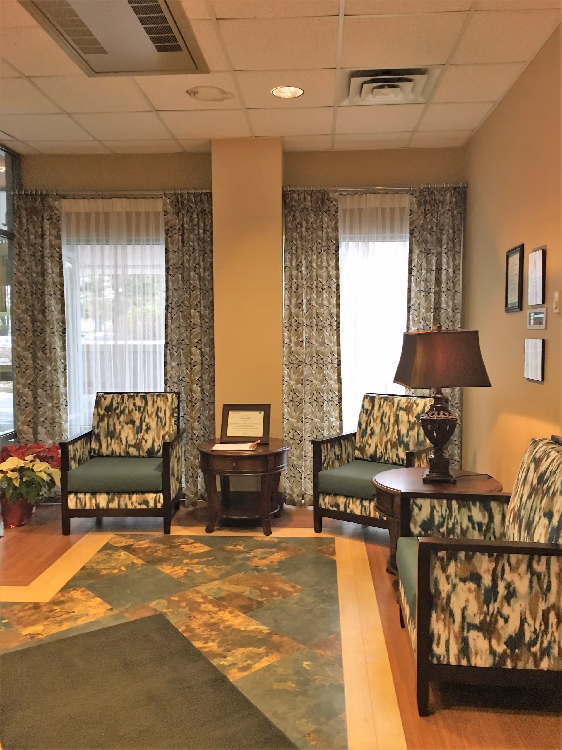 Small lobby seating area with patterned armchairs, side tables, a lamp, and curtained windows.