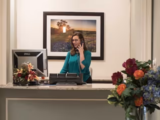 A woman standing behind a reception desk talking on the phone. There is a Dell computer monitor on the desk and a framed landscape photograph on the wall behind her. A bouquet of flowers is visible in the foreground on the right side.