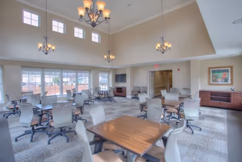 A spacious, well-lit common area with multiple tables and chairs arranged for group seating. The room features high ceilings with hanging chandeliers, large windows allowing natural light, and neutral-colored walls. There is a wooden cabinet with a framed picture on the right side and an open doorway leading to another room.