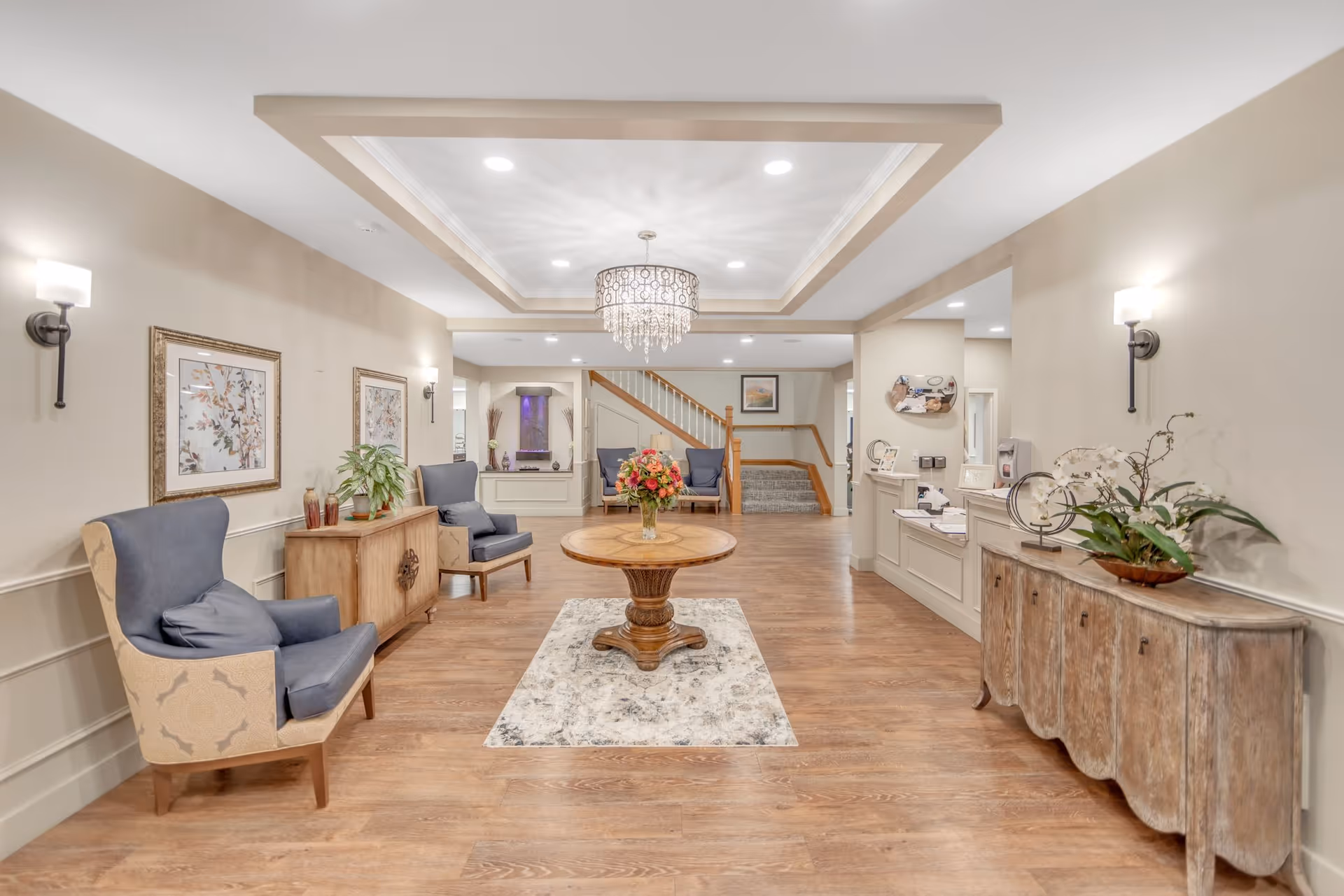A spacious and well-lit interior common area with wooden flooring, beige walls, and a decorative ceiling light fixture. The room features a round wooden table with a floral arrangement in the center on a patterned rug, two blue cushioned armchairs with wooden frames on the left, and a wooden sideboard with plants and decorative items on the right. In the background, there is a staircase with wooden railings and additional seating with two blue chairs. Wall sconces provide additional lighting, and framed artwork decorates the walls.