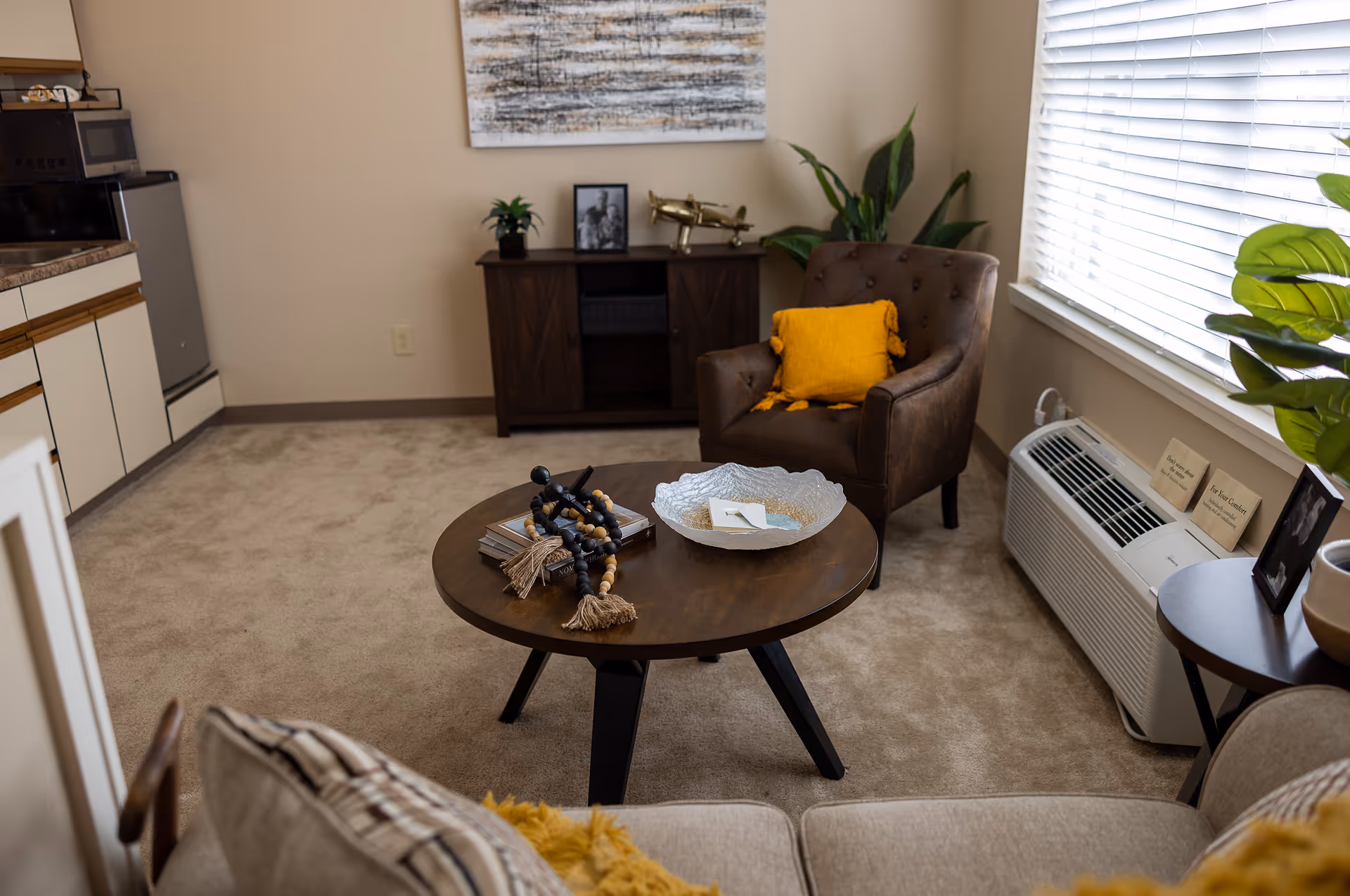 Cozy living room with a round wooden coffee table, armchair with a yellow pillow, sofa, and a small kitchenette area.