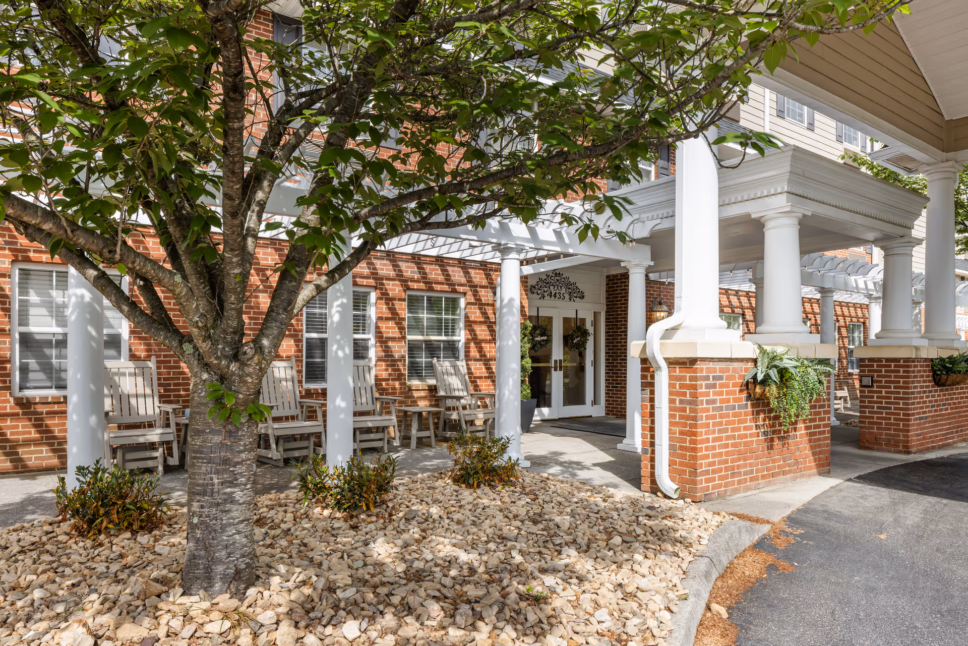 Front entrance of a brick senior living facility with white columns, a covered portico, rocking chairs and a tree in a landscaped bed.