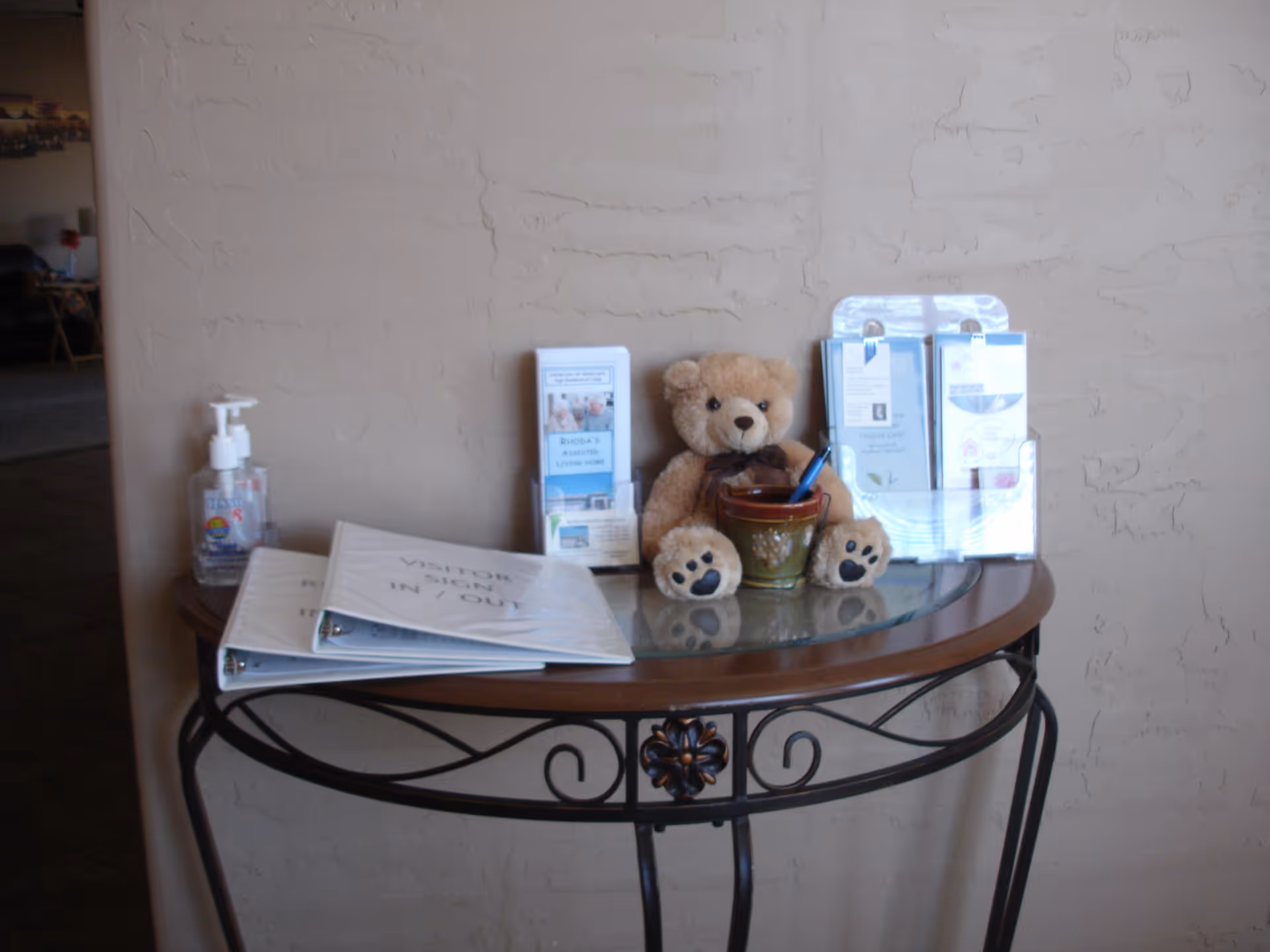A small decorative table against a textured beige wall with a visitor sign-in/out binder, a bottle of hand sanitizer, informational brochures, a teddy bear, and a small container holding a pen.