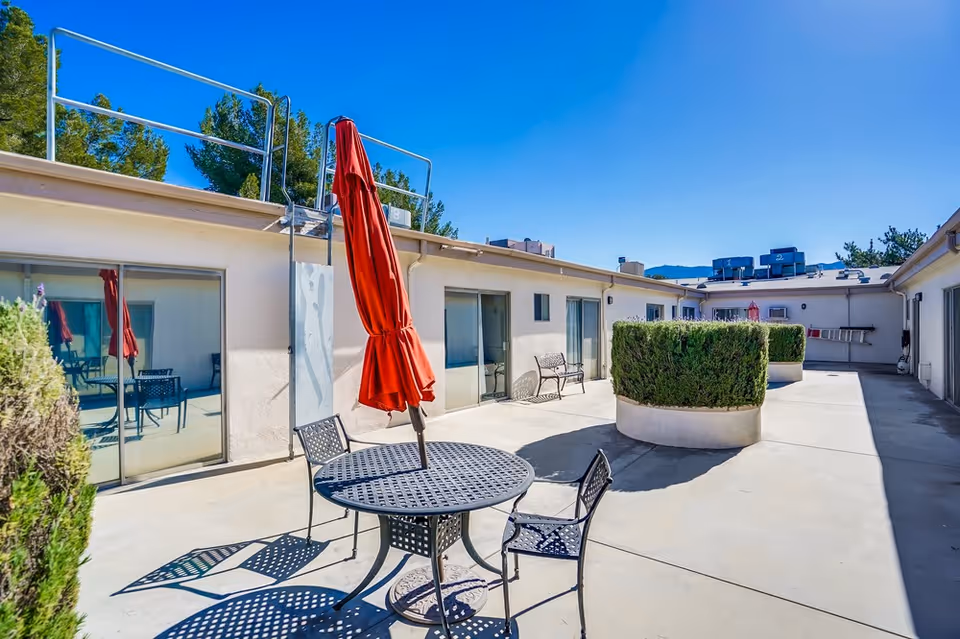 Outdoor courtyard area at Santa Clarita Post Acute with a round metal table and three chairs, a closed red umbrella, potted bushes, sliding glass doors leading to interior rooms, and a clear blue sky.