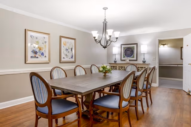 A dining room with a long wooden table surrounded by eight wooden chairs with blue cushioned seats and striped backs. The room has light beige walls with white trim, two framed floral artworks on one wall, a chandelier hanging above the table, and a sideboard with two lamps and a framed decorative piece. There is an open doorway leading to another room.