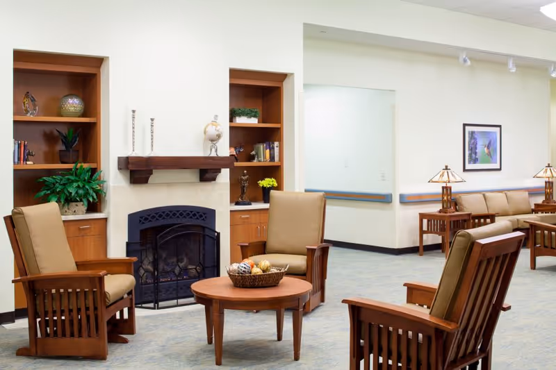 A cozy seating area in a senior living facility featuring four wooden armchairs with beige cushions arranged around a round wooden coffee table with a decorative basket. Behind the chairs is a fireplace with a wooden mantel, flanked by built-in shelves holding plants, books, and decorative items. In the background, there is a beige sofa with side tables and lamps, and a framed picture on the wall.