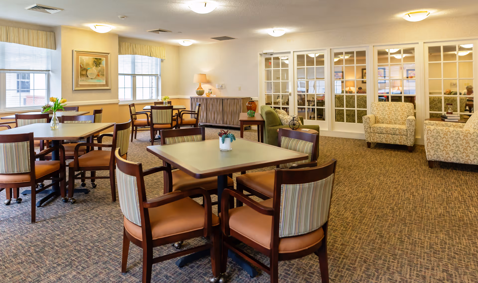 A well-lit common area with several square tables surrounded by wooden chairs with striped cushions. There are two armchairs with leaf-patterned upholstery and a green sofa near a wooden side table with decorative items. Large windows with blinds and valances let in natural light. A set of glass-paneled double doors leads to another room with more seating and people visible inside.