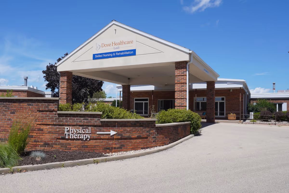 Exterior view of Dove Healthcare - Lodi facility entrance with a covered drop-off area supported by brick pillars. A brick wall in front has a sign pointing to Physical Therapy. The sky is clear and blue.