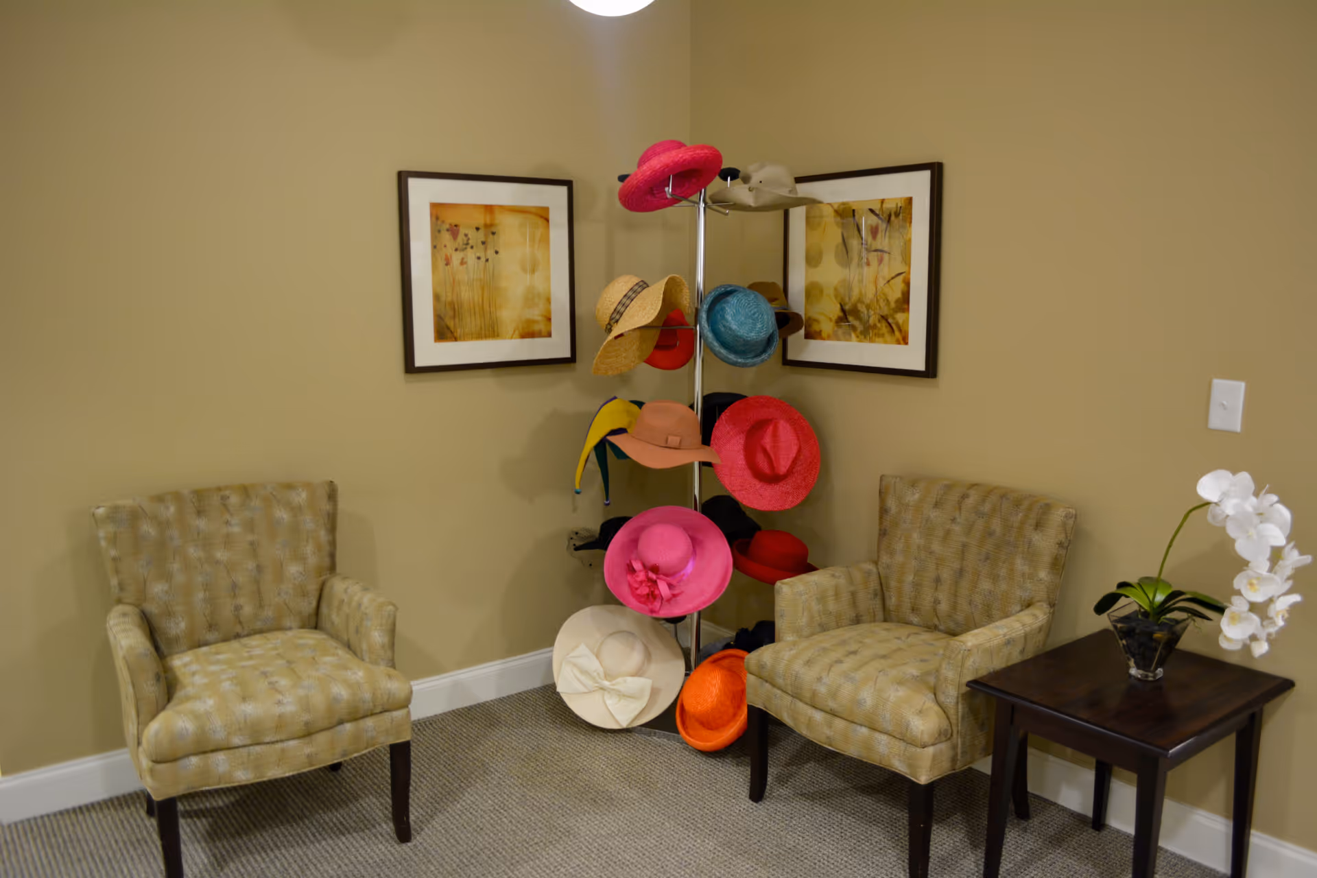 A cozy corner with two beige patterned armchairs facing each other, a small dark wooden side table with a white orchid plant, and a metal hat stand holding various colorful hats. Two framed abstract floral paintings hang on the beige walls behind the hat stand.