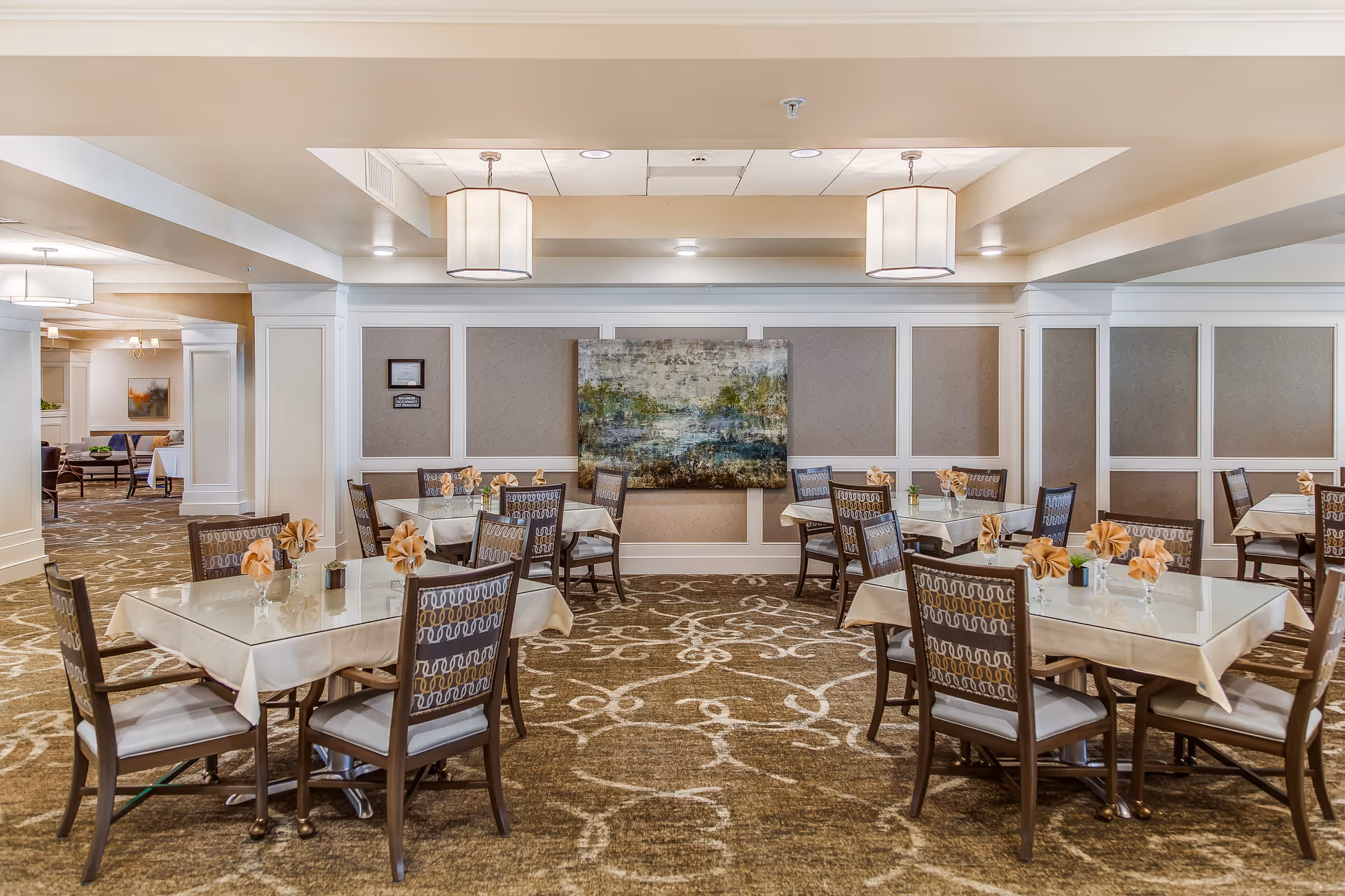 A well-lit dining room with several square tables covered with white tablecloths and glass tops. Each table is surrounded by wooden chairs with patterned upholstery. The tables are decorated with folded beige napkins and small plants. The room has beige walls with white paneling and a large abstract painting on the far wall. The ceiling features recessed lighting and two large hanging light fixtures.