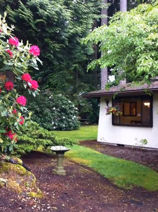 A lush garden area with green grass, flowering bushes with pink and white flowers, and tall trees surrounding a small white building with a window and hanging flower basket.