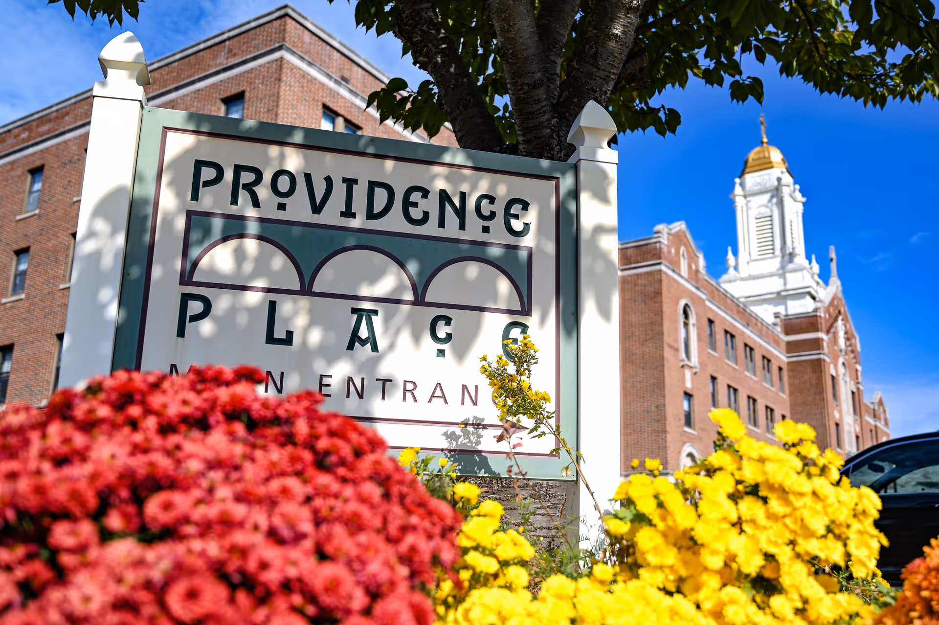 A sign reading Providence Place by colorful flowers with a large brick building and white cupola in the background.
