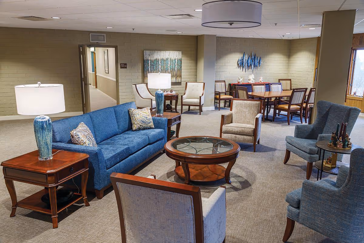 Spacious senior living common area with a blue sofa, several armchairs, coffee tables, lamps, and a dining table in the background.