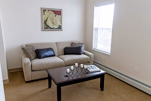 Small living room with a beige loveseat, decorative pillows, a dark wood coffee table with candles and a book, and a window with blinds.