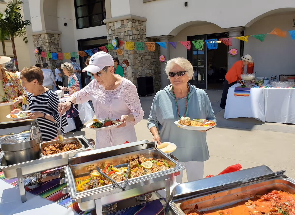 Two elderly women serving themselves food from buffet trays at an outdoor event. One woman is wearing a white cap and light pink top, while the other is wearing sunglasses and a light blue top. Colorful paper decorations hang above, and other people are visible in the background near a building entrance.