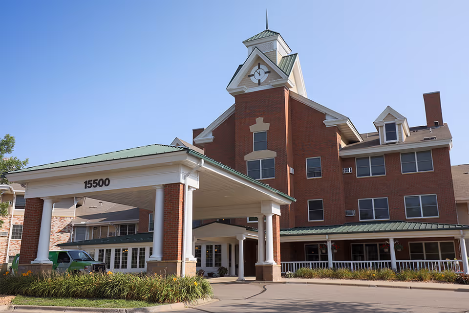 Exterior view of Plymouth Towne Square senior living facility showing a large brick building with multiple windows, a green metal roof, and a covered entrance with white columns and the number 15500 displayed. There is a driveway in front and some landscaping with plants and flowers.