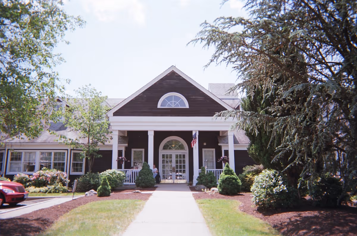 Front exterior view of a senior living facility named Blessed House with a paved walkway leading to the entrance. The building has a dark brown facade with white trim, large windows, and a covered porch supported by white columns. There are trees, shrubs, and landscaped garden beds on either side of the walkway, and an American flag is displayed near the entrance.