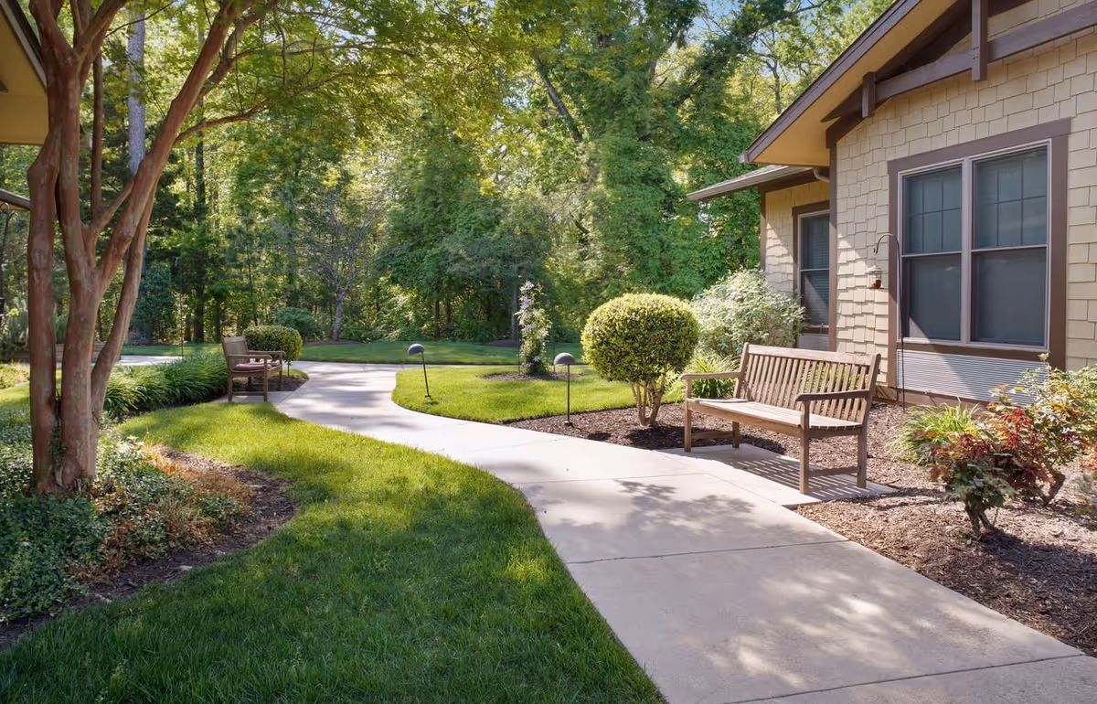 A peaceful outdoor garden area with a curved concrete pathway, wooden benches, manicured bushes, trees, and a building with beige siding and brown trim in the background.
