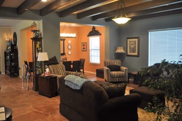 A cozy living room area with brown sofas and armchairs, a side table with a lamp, and a large window with blinds. The room has exposed wooden beams on the ceiling and a warm color palette. In the background, there is a dining area with a table and chairs, and decorative wall art.