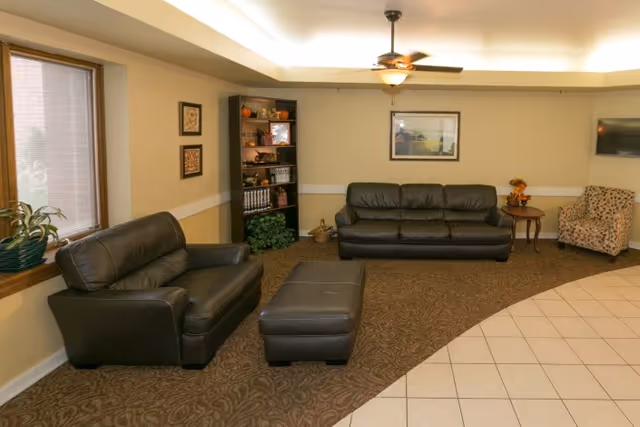 A cozy common room with leather sofas, an ottoman, an armchair, a bookcase, and a tiled entryway under recessed lighting.