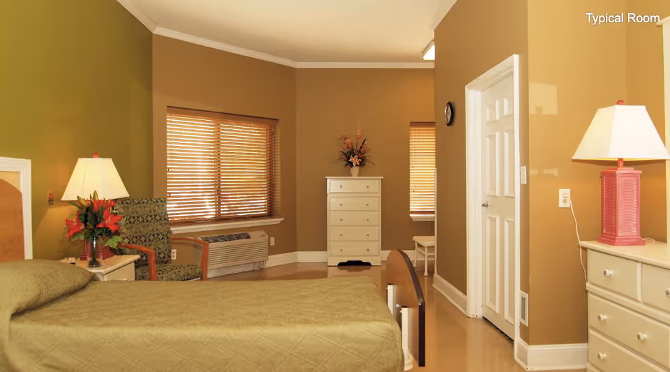 A typical bedroom in a senior living facility with a single bed covered in a green quilt, a nightstand with a lamp and flowers, a patterned armchair, two windows with wooden blinds, a white dresser with a flower vase on top, and a door leading to another room.