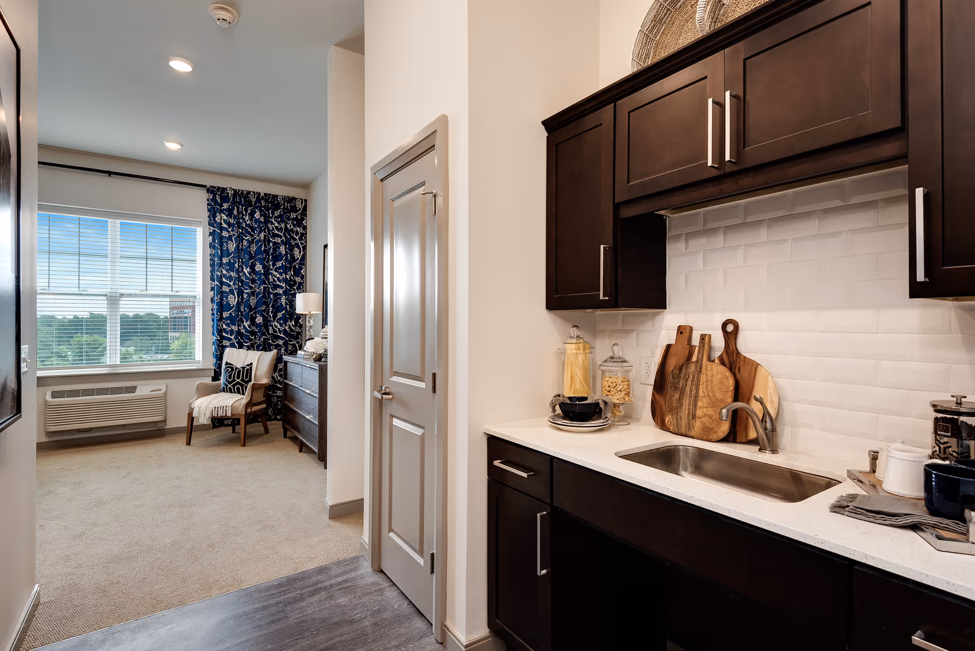 Interior view of a senior living facility showing a kitchenette with dark wood cabinets, a white countertop, a small sink, and decorative cutting boards. To the left, there is a doorway leading to a room with a large window, blue patterned curtains, a chair with a pillow and throw, and a dresser with a lamp.