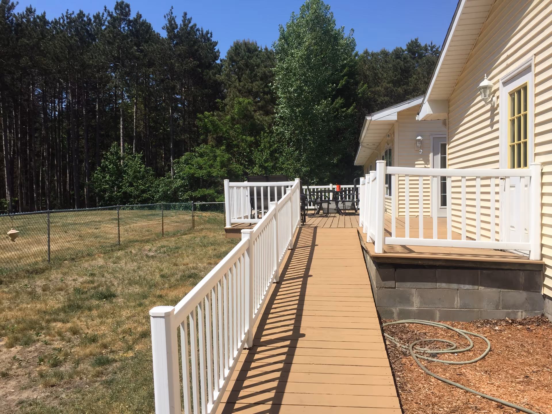Outdoor view of a wooden ramp with white railings leading to a yellow building with a deck area that has a table and chairs. The area is surrounded by a grassy yard and a chain-link fence, with tall trees in the background under a clear blue sky.
