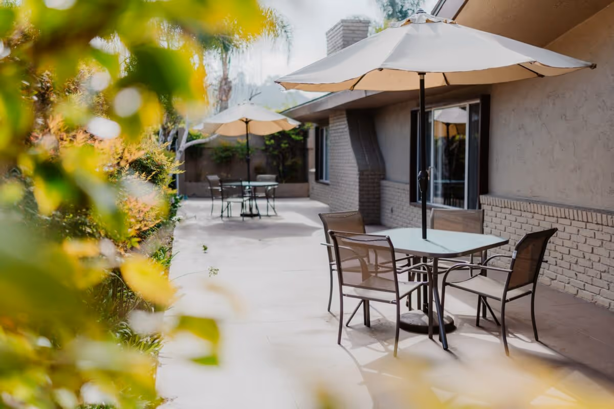 Outdoor patio area with tables and chairs under large umbrellas next to a building with brick and stucco walls, surrounded by greenery and palm trees in the background.