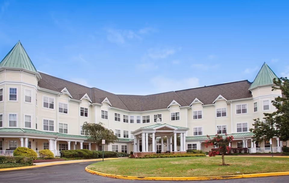 Exterior front view of a three-story pale-yellow senior living building with green-roofed turrets, a covered columned entrance, and a landscaped circular drive.