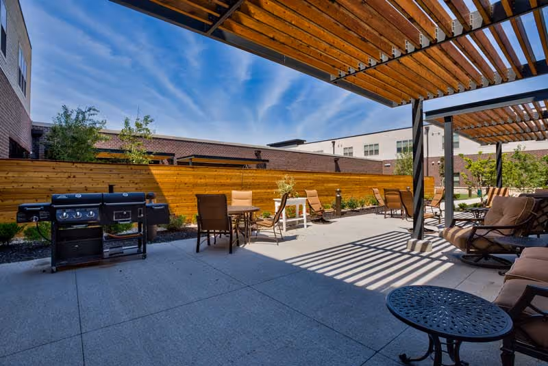 Outdoor patio courtyard with pergola, seating areas, tables, and a barbecue grill at a senior living facility.