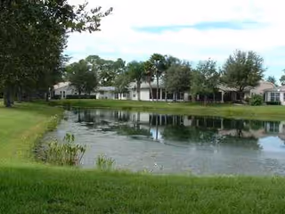 A pond with grassy shores reflects a row of senior living buildings and trees.