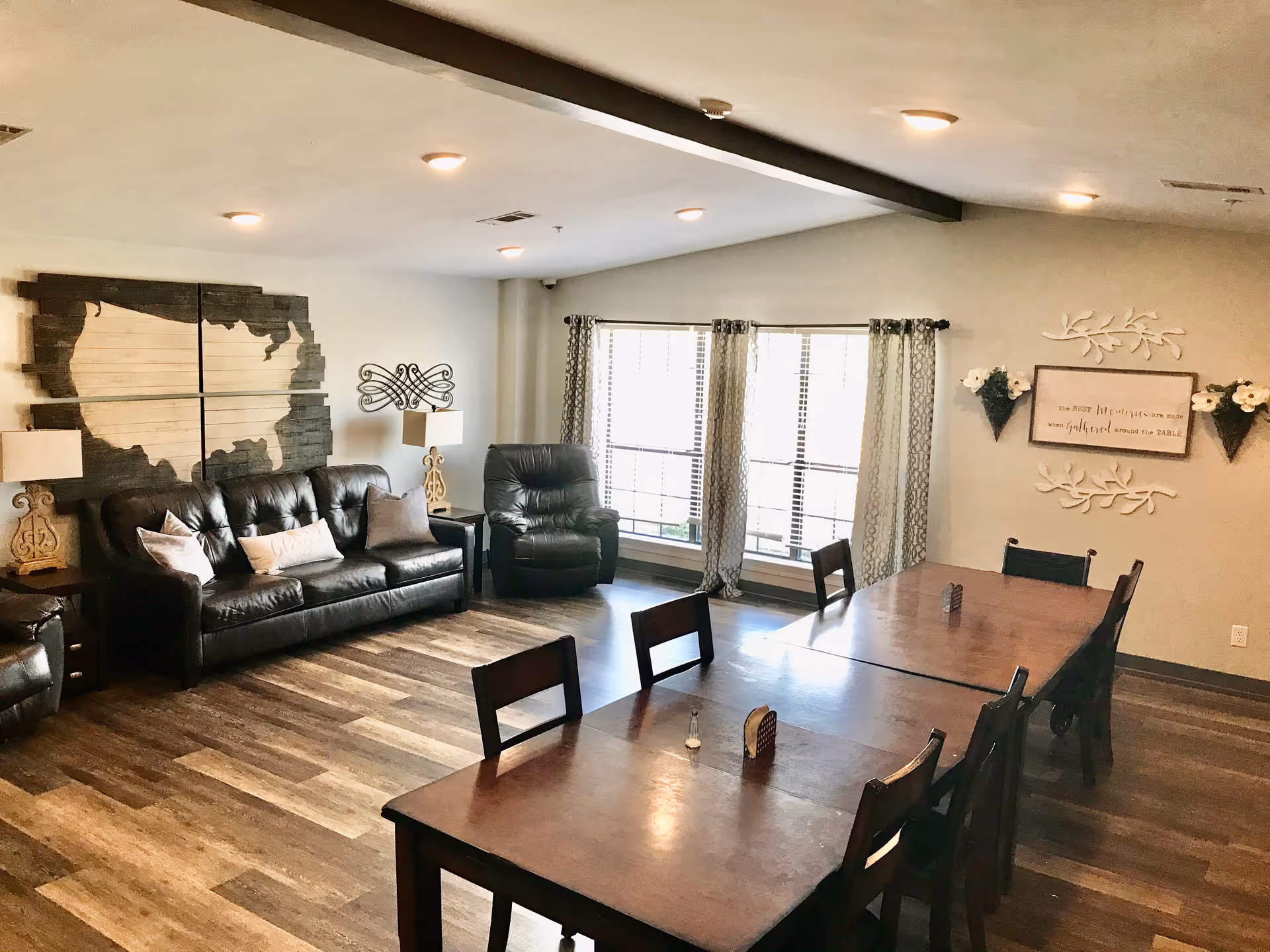 Bright communal living and dining room with leather sofas and a recliner, long wooden tables and decorative wall art including a wooden map of the U.S.
