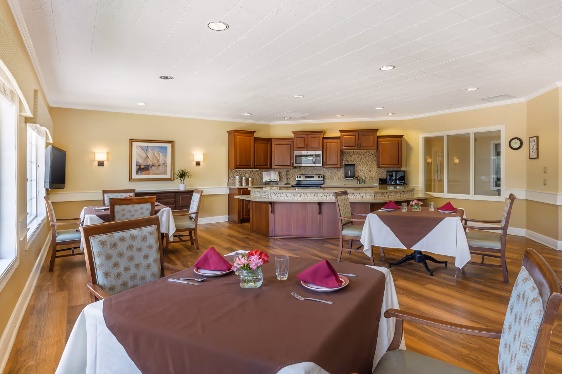 A dining area in a senior living facility with several tables covered with white and brown tablecloths, each set with plates, glasses, silverware, and folded red napkins. The room has wooden flooring, beige walls, and a kitchen area with wooden cabinets, a stove, microwave, and coffee maker in the background. There is a framed picture on the wall and a flat-screen TV mounted near the windows on the left side.