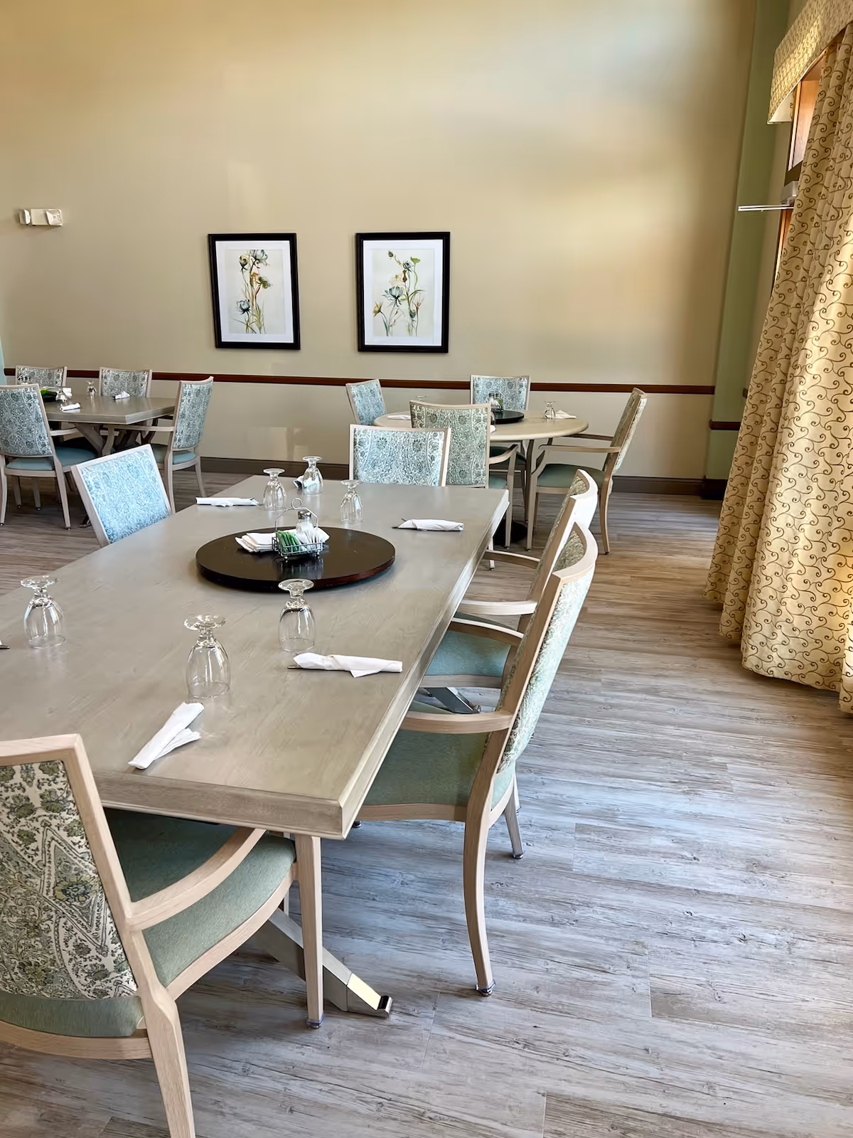 A dining room with several tables and chairs. The tables are set with upside-down glasses and folded napkins. The chairs have light green upholstery with a floral pattern. Two framed floral artworks hang on the beige wall. A large window with patterned curtains is on the right side, letting in natural light. The floor is light wood.
