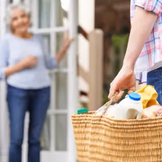 A person carries a wicker basket of groceries toward the open doorway of a senior living facility, with an older woman standing in the background.