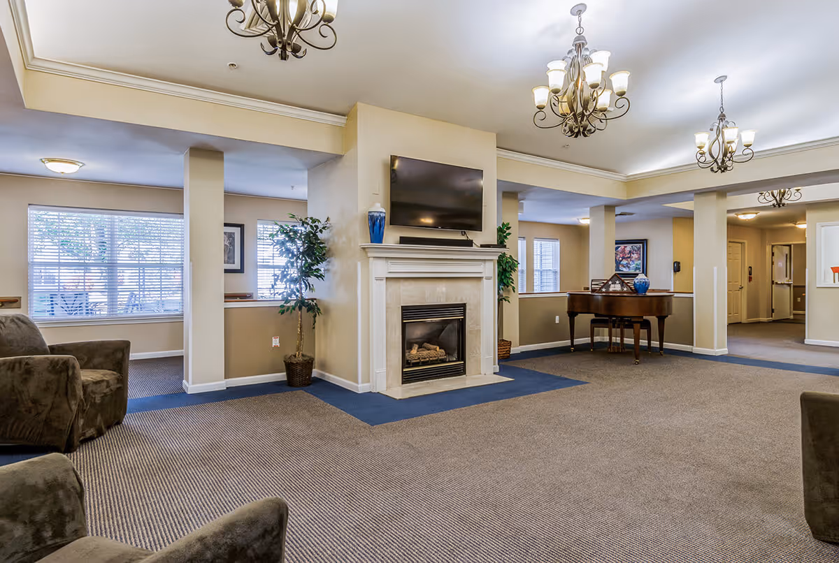 Spacious living room area in a senior living facility with a fireplace and a mounted flat-screen TV above it. The room features multiple chandeliers, large windows with blinds, a grand piano, potted plants, and comfortable armchairs arranged around the space. The walls are painted in neutral tones and the floor is carpeted.