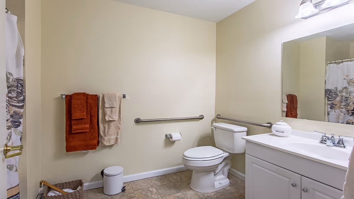 A clean bathroom with beige walls and tiled floor. There is a white toilet with metal grab bars on both sides, a white vanity with a sink and a large mirror above it. On the left wall, there are two towel racks holding brown and beige towels. A small white trash can and a woven basket with rolled towels are on the floor. A shower curtain with a floral pattern is partially visible on the left side.