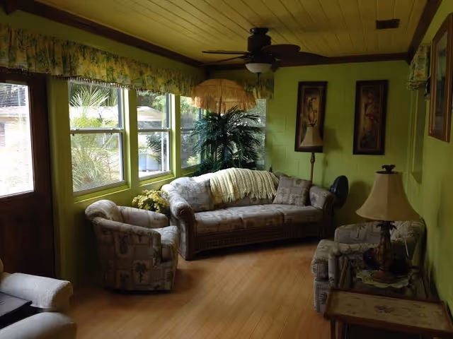 Sunlit living room with bright green walls, a sofa and two armchairs, multiple windows, potted plants, lamps, and a ceiling fan.