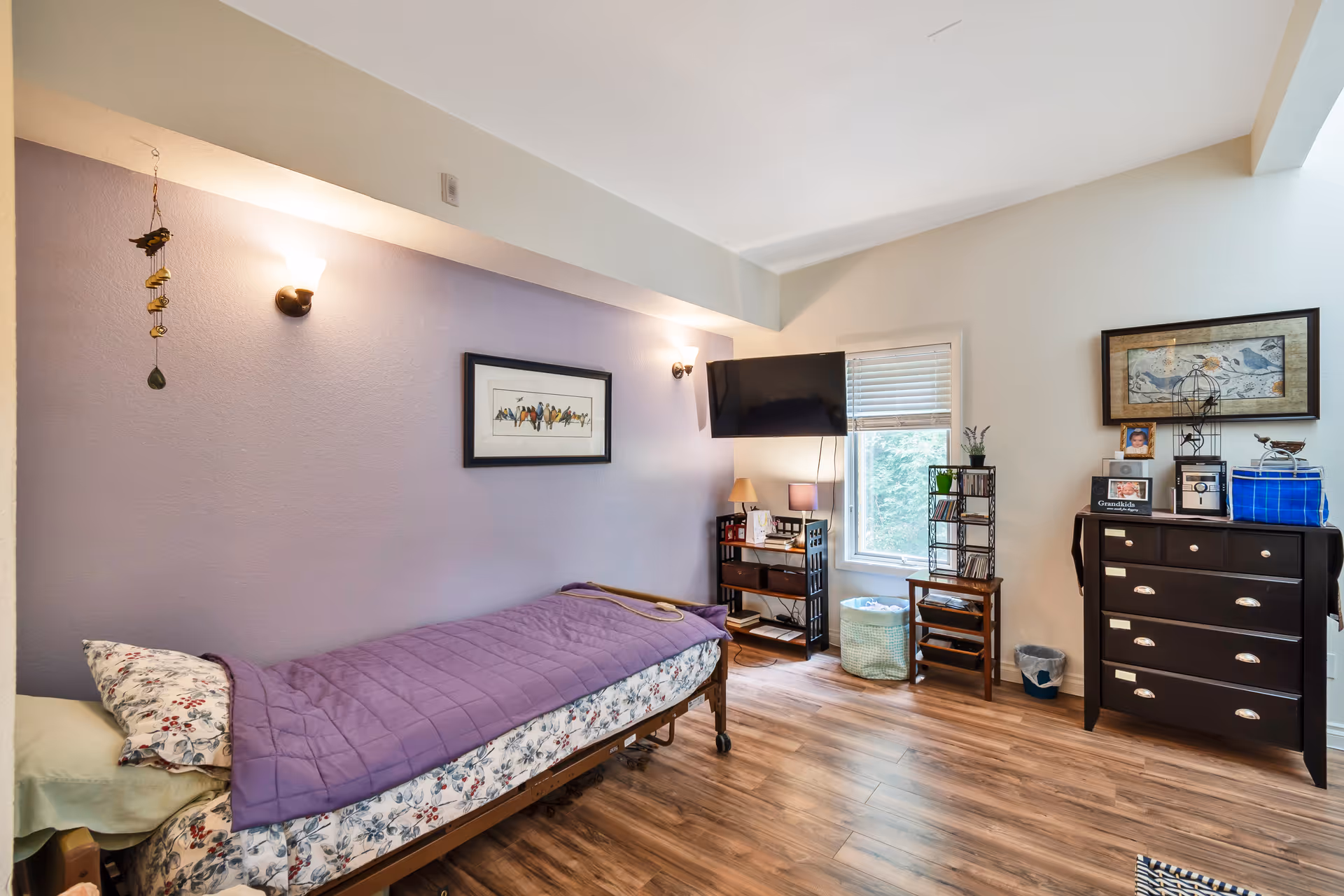 A cozy bedroom in a senior living facility with a single bed covered with a purple quilt and floral sheets. The room has light purple and white walls, wooden flooring, a wall-mounted TV, a window with blinds, a black dresser with framed photos and decorations, and a small shelving unit with a lamp and other items.