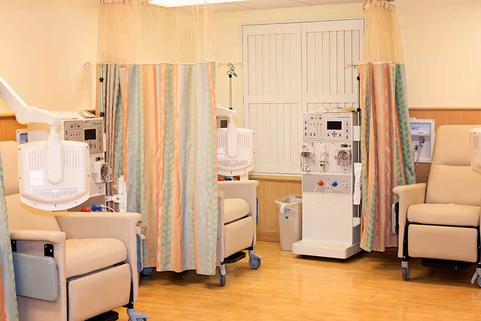 A medical treatment room with beige reclining chairs separated by colorful privacy curtains. Each chair is equipped with medical devices, including dialysis machines and monitors. The room has wooden flooring and a window with closed shutters.