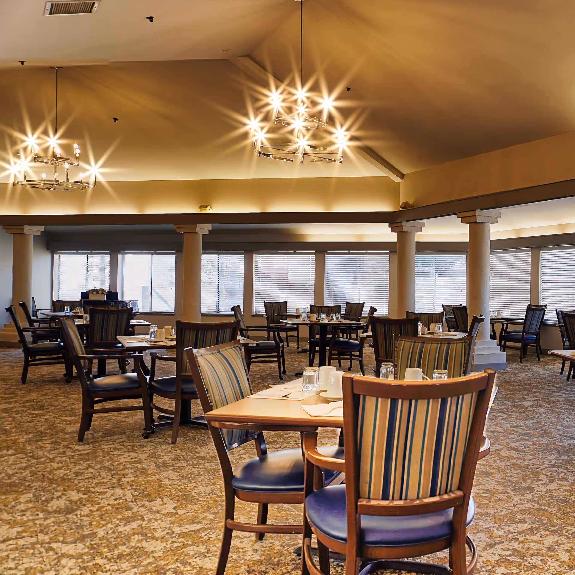 A spacious dining room with multiple wooden tables and chairs arranged neatly. The chairs have striped upholstery and the tables are set with glasses, cups, and napkins. The room features large windows with blinds, carpeted floors, and elegant chandeliers hanging from a high ceiling.