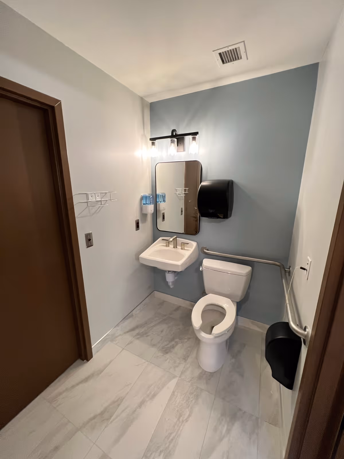 Accessible bathroom with a toilet (open-front seat), wall-mounted sink and mirror, grab bars, soap and paper dispensers, and tiled floor.
