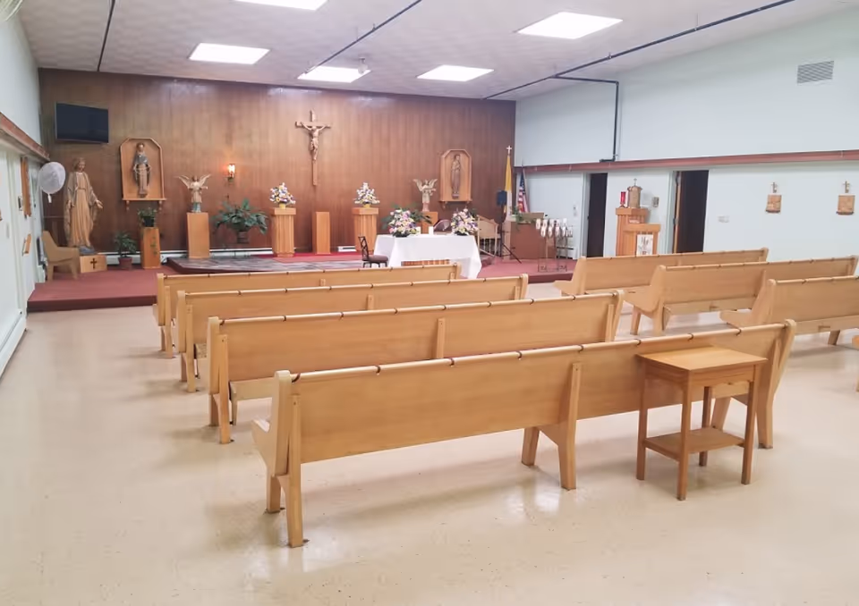 Interior view of a chapel with wooden pews arranged in rows facing a raised platform. The platform has religious statues, floral arrangements, a crucifix on the wooden-paneled wall, and a table covered with a white cloth. The room has a light-colored floor and ceiling with fluorescent lights.