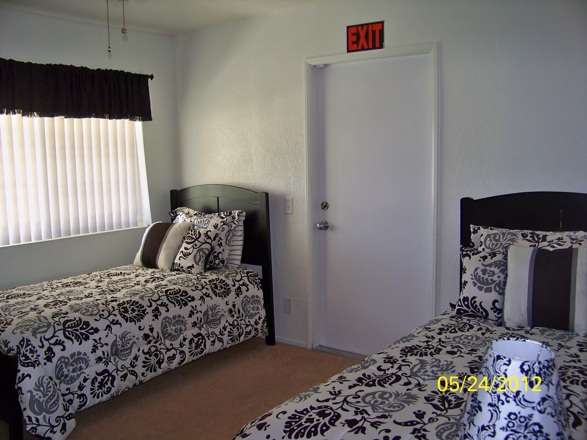 A bedroom with two single beds, each with black and white patterned bedding and multiple pillows. There is a window with vertical blinds and a black valance on the left wall. A white door with a red EXIT sign above it is on the far wall. The carpet is beige, and the walls are white.